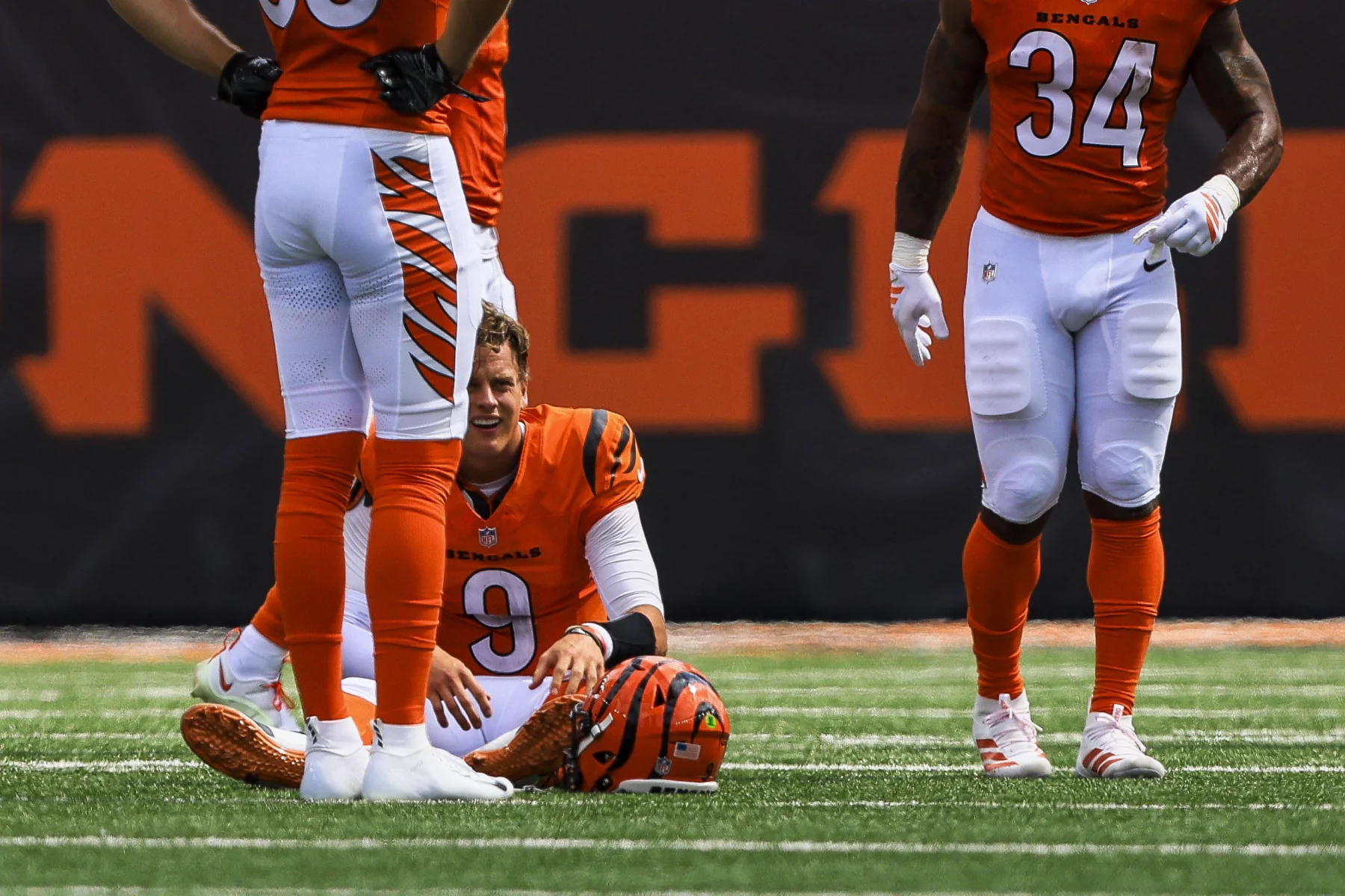 NFL Spieltag 2 - Joe Burrow im Bengals-Trikot mit der Nummer 9 sitzt nach dem NFL-Spieltag 2 auf dem Spielfeld und sieht verzweifelt aus. Sein Helm liegt auf dem Boden, während zwei orange-weiß gekleidete Mannschaftskameraden die Sicht teilweise versperren, wobei der Rasen und ein großes oranges Schild zu sehen sind. Diese Beschreibung wurde mit der FootballR KI automatisch generiert.