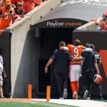 Ein Football-Spieler in einem orangefarbenen Trikot mit der Nummer 9, bei dem es sich vermutlich um Joe Burrow verletzt handelt, wird von Mitarbeitern unterstützt, während er auf einen Stadiontunnel mit der Aufschrift Paycor Stadium" zugeht, während Fotografen und Fans in orangefarbener Kleidung zuschauen. Diese Beschreibung wurde mit der FootballR KI automatisch generiert.