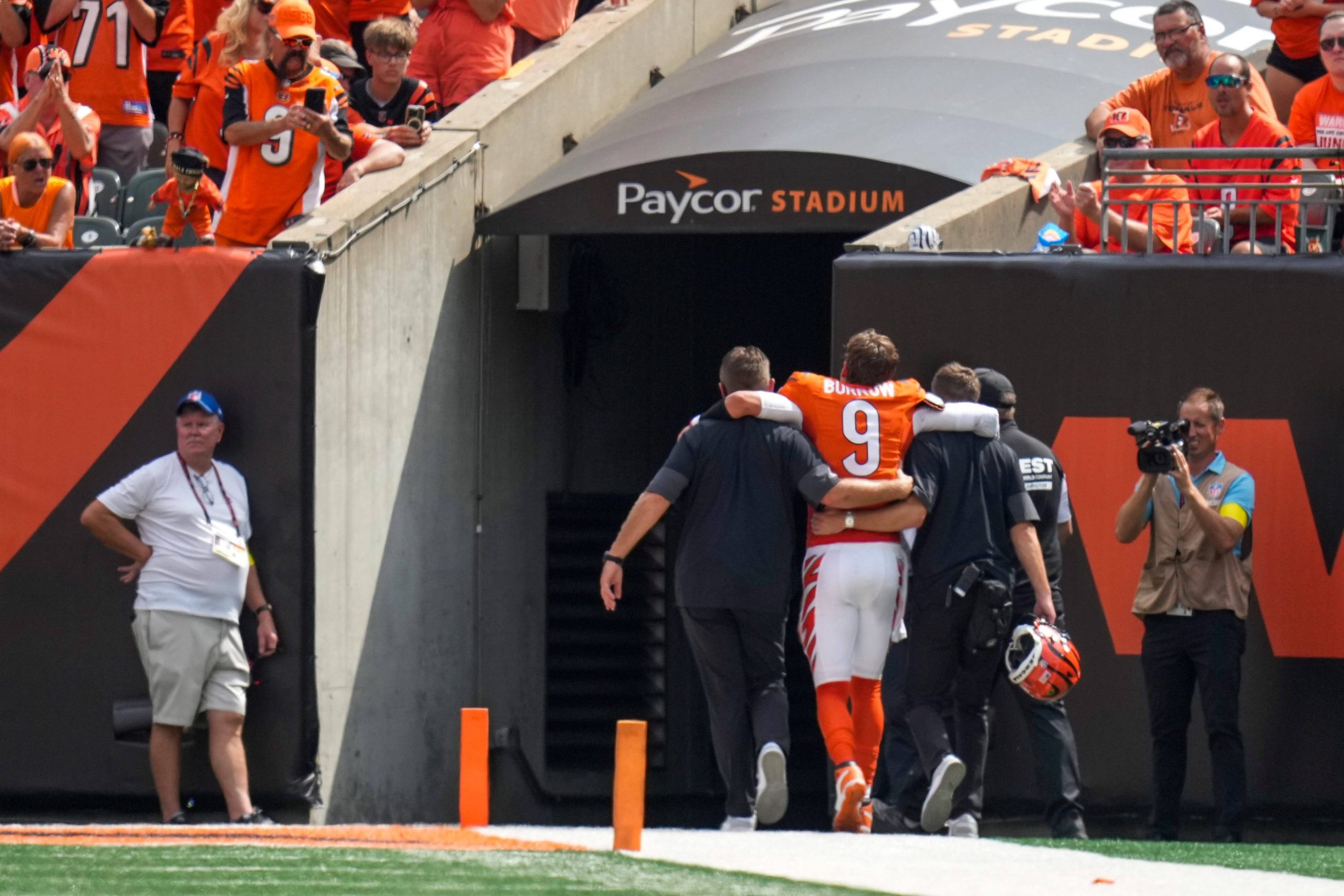 Ein Football-Spieler in einem orangefarbenen Trikot mit der Nummer 9, bei dem es sich vermutlich um Joe Burrow verletzt handelt, wird von Mitarbeitern unterstützt, während er auf einen Stadiontunnel mit der Aufschrift Paycor Stadium" zugeht, während Fotografen und Fans in orangefarbener Kleidung zuschauen. Diese Beschreibung wurde mit der FootballR KI automatisch generiert.