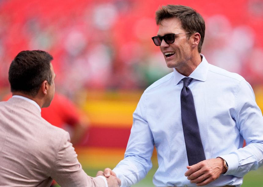 Tom Brady mit weißem Hemd, dunkler Krawatte und Sonnenbrille lächelt breit, während er einem anderen Mann in einem hellbraunen Anzug die Hand schüttelt. Das unscharfe Stadion deutet auf eine Sportveranstaltung hin, bei der vielleicht Raiders-Miteigentümer Tom Brady im Rampenlicht steht. Diese Beschreibung wurde mit der FootballR KI automatisch generiert.