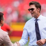 Tom Brady mit weißem Hemd, dunkler Krawatte und Sonnenbrille lächelt breit, während er einem anderen Mann in einem hellbraunen Anzug die Hand schüttelt. Das unscharfe Stadion deutet auf eine Sportveranstaltung hin, bei der vielleicht Raiders-Miteigentümer Tom Brady im Rampenlicht steht. Diese Beschreibung wurde mit der FootballR KI automatisch generiert.