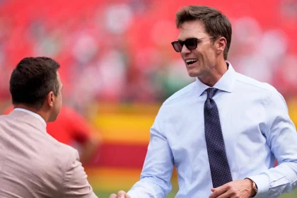 Tom Brady mit weißem Hemd, dunkler Krawatte und Sonnenbrille lächelt breit, während er einem anderen Mann in einem hellbraunen Anzug die Hand schüttelt. Das unscharfe Stadion deutet auf eine Sportveranstaltung hin, bei der vielleicht Raiders-Miteigentümer Tom Brady im Rampenlicht steht. Diese Beschreibung wurde mit der FootballR KI automatisch generiert.