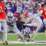 Ein Kicker in einer weiß-blauen Uniform schießt den Ball, während ein Halter auf dem Feld kniet. Denver Broncos Verteidiger in orangefarbenen Trikots stürmen auf sie zu. Die Fans im Stadion füllen den unscharfen Hintergrund und beobachten die intensive Aktion. Diese Beschreibung wurde mit der FootballR KI automatisch generiert.