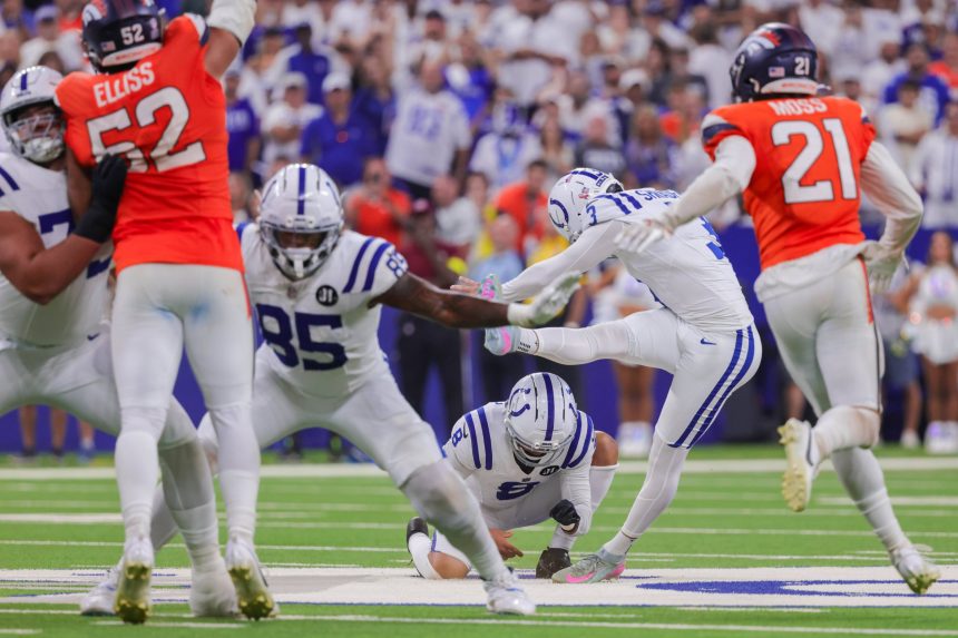 Ein Kicker in einer weiß-blauen Uniform schießt den Ball, während ein Halter auf dem Feld kniet. Denver Broncos Verteidiger in orangefarbenen Trikots stürmen auf sie zu. Die Fans im Stadion füllen den unscharfen Hintergrund und beobachten die intensive Aktion. Diese Beschreibung wurde mit der FootballR KI automatisch generiert.