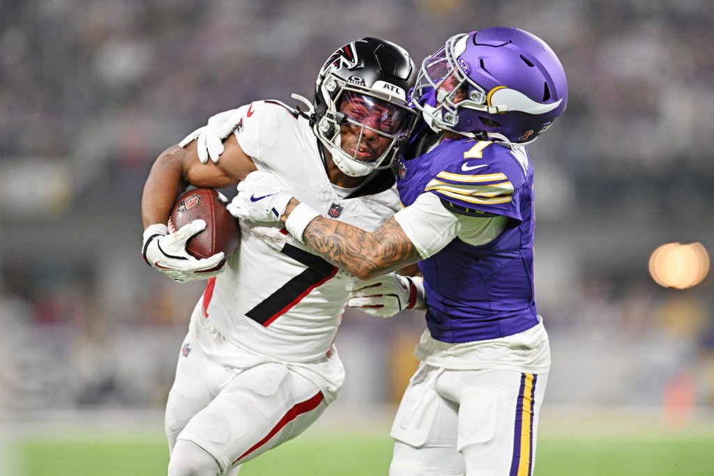 Der Running Back der Atlanta Falcons, Bijan Robinson, versucht sich zu befreien, als ihn ein Verteidiger der Minnesota Vikings in einem lila Trikot bei einem Tackling-Versuch auf einem gut beleuchteten Football-Feld umschließt. Diese Beschreibung wurde mit der FootballR KI automatisch generiert.