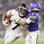 Der Running Back der Atlanta Falcons, Bijan Robinson, versucht sich zu befreien, als ihn ein Verteidiger der Minnesota Vikings in einem lila Trikot bei einem Tackling-Versuch auf einem gut beleuchteten Football-Feld umschließt. Diese Beschreibung wurde mit der FootballR KI automatisch generiert.