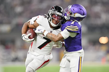 Der Running Back der Atlanta Falcons, Bijan Robinson, versucht sich zu befreien, als ihn ein Verteidiger der Minnesota Vikings in einem lila Trikot bei einem Tackling-Versuch auf einem gut beleuchteten Football-Feld umschließt. Diese Beschreibung wurde mit der FootballR KI automatisch generiert.