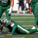 Justin Fields in einem grünen Trikot und mit Helm der New York Jets stemmt sich während eines Spiels auf Händen und Knien vom Boden hoch, während zwei Mannschaftskameraden daneben stehen. Auf dem sonnenbeschienenen Spielfeld herrscht eine Energie, die an ein Highlight von Justin Fields erinnert. Diese Beschreibung wurde mit der FootballR KI automatisch generiert.