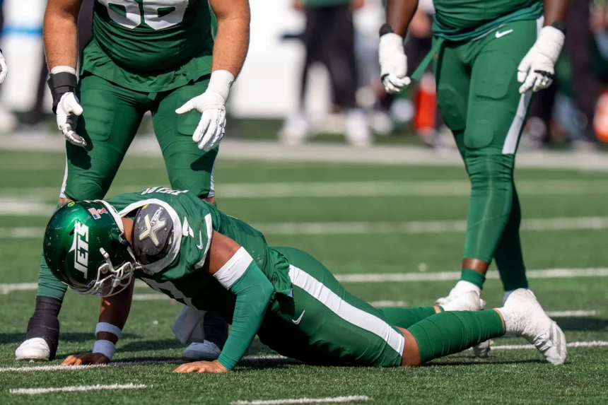 Justin Fields in einem grünen Trikot und mit Helm der New York Jets stemmt sich während eines Spiels auf Händen und Knien vom Boden hoch, während zwei Mannschaftskameraden daneben stehen. Auf dem sonnenbeschienenen Spielfeld herrscht eine Energie, die an ein Highlight von Justin Fields erinnert. Diese Beschreibung wurde mit der FootballR KI automatisch generiert.