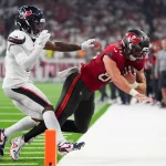 Ein Footballspieler in einem roten Trikot der Tampa Bay Buccaneers springt mit dem Ball in Richtung Endzone, dicht verfolgt von C.J. Gardner-Johnson. Die Aktion findet in der Nähe der Seitenlinie statt, wobei der orangefarbene Pylon die Torlinie markiert. Diese Beschreibung wurde mit der FootballR KI automatisch generiert.