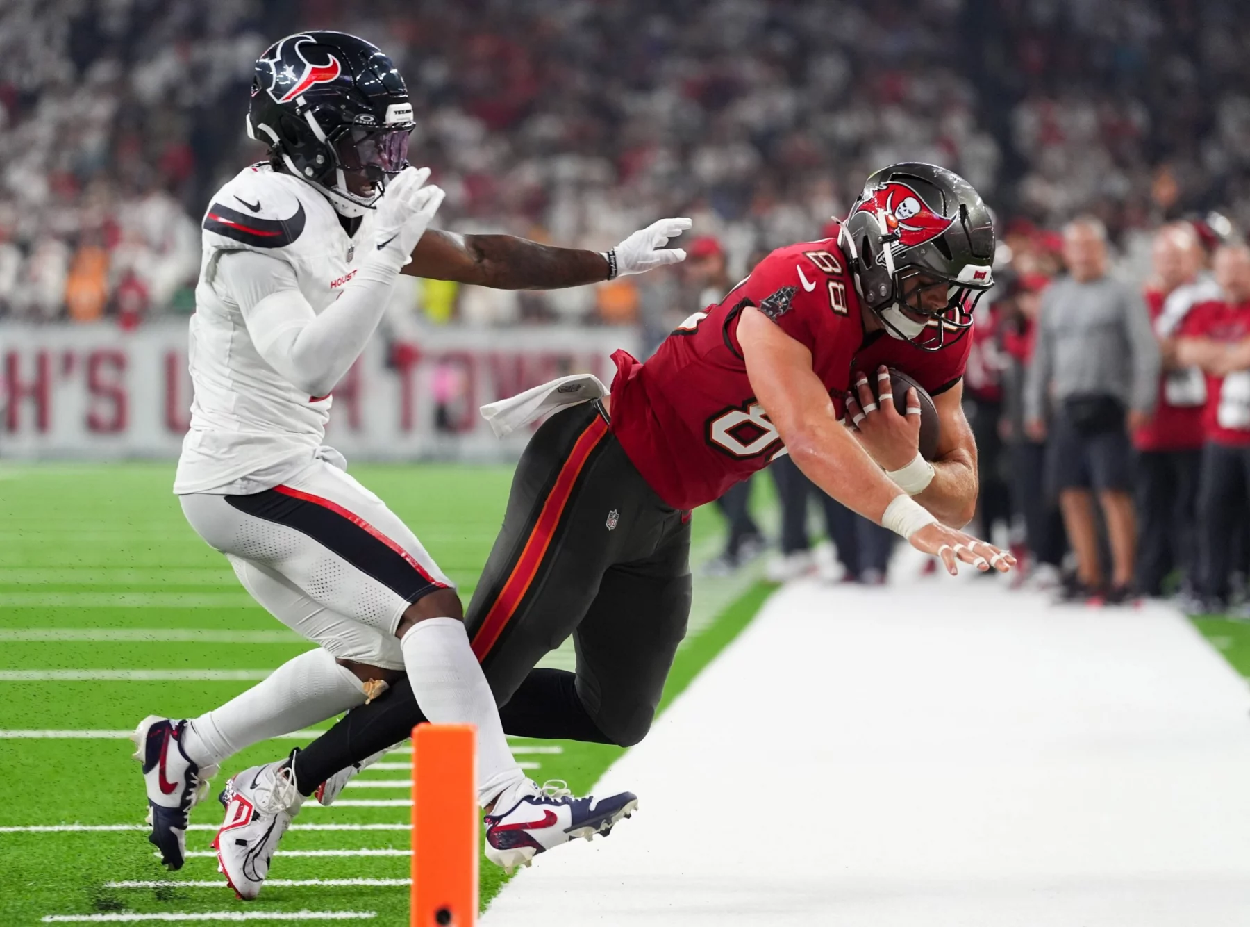 Ein Footballspieler in einem roten Trikot der Tampa Bay Buccaneers springt mit dem Ball in Richtung Endzone, dicht verfolgt von C.J. Gardner-Johnson. Die Aktion findet in der Nähe der Seitenlinie statt, wobei der orangefarbene Pylon die Torlinie markiert. Diese Beschreibung wurde mit der FootballR KI automatisch generiert.