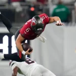 Ein Football-Spieler in einem roten Trikot, der Baker Mayfield ähnelt, ist in der Luft, während er den Ball umklammert und von einem Spieler in Weiß auf einem grünen Feld angegriffen wird. Im Hintergrund sind unscharfe Figuren und ein Teil einer digitalen Anzeigetafel zu sehen. Diese Beschreibung wurde mit der FootballR KI automatisch generiert.