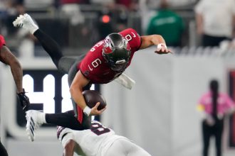Ein Football-Spieler in einem roten Trikot, der Baker Mayfield ähnelt, ist in der Luft, während er den Ball umklammert und von einem Spieler in Weiß auf einem grünen Feld angegriffen wird. Im Hintergrund sind unscharfe Figuren und ein Teil einer digitalen Anzeigetafel zu sehen. Diese Beschreibung wurde mit der FootballR KI automatisch generiert.