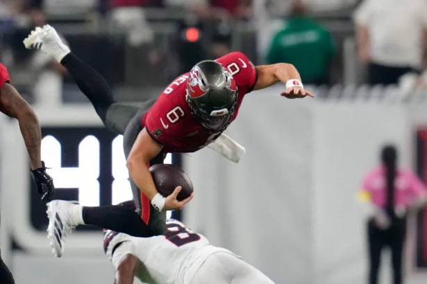 Ein Football-Spieler in einem roten Trikot, der Baker Mayfield ähnelt, ist in der Luft, während er den Ball umklammert und von einem Spieler in Weiß auf einem grünen Feld angegriffen wird. Im Hintergrund sind unscharfe Figuren und ein Teil einer digitalen Anzeigetafel zu sehen. Diese Beschreibung wurde mit der FootballR KI automatisch generiert.