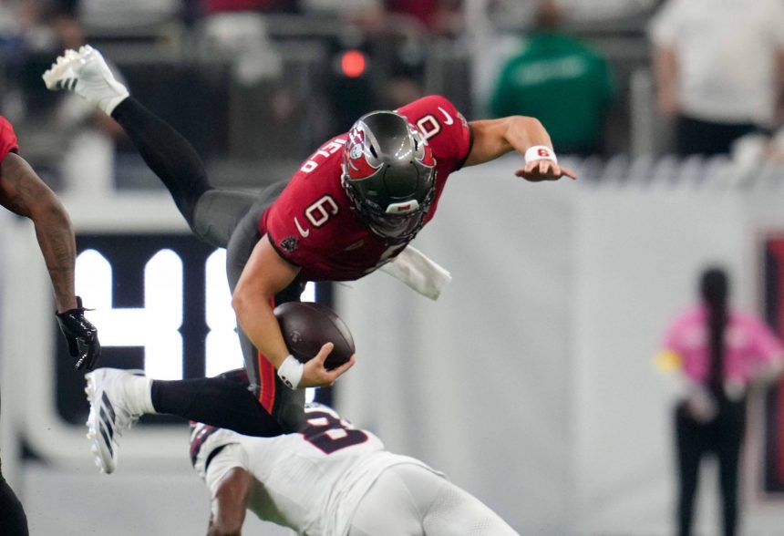 Ein Football-Spieler in einem roten Trikot, der Baker Mayfield ähnelt, ist in der Luft, während er den Ball umklammert und von einem Spieler in Weiß auf einem grünen Feld angegriffen wird. Im Hintergrund sind unscharfe Figuren und ein Teil einer digitalen Anzeigetafel zu sehen. Diese Beschreibung wurde mit der FootballR KI automatisch generiert.