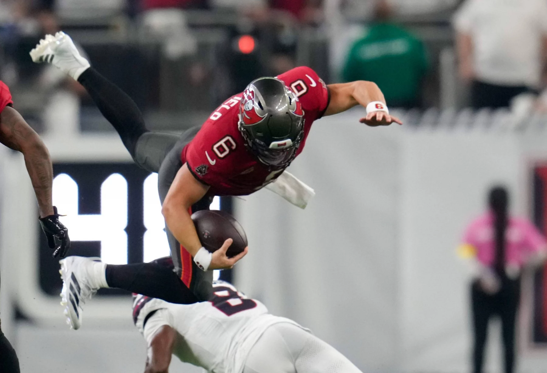 Ein Football-Spieler in einem roten Trikot, der Baker Mayfield ähnelt, ist in der Luft, während er den Ball umklammert und von einem Spieler in Weiß auf einem grünen Feld angegriffen wird. Im Hintergrund sind unscharfe Figuren und ein Teil einer digitalen Anzeigetafel zu sehen. Diese Beschreibung wurde mit der FootballR KI automatisch generiert.