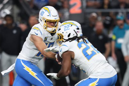 Ein Footballspieler im weißen Chargers-Trikot mit der Nummer 10 übergibt den Ball während eines Spiels an seinen Teamkollegen Najee Harris. Beide tragen Helme und sind fokussiert, mit unscharfen Zuschauern und einer digitalen Nummernanzeige im Hintergrund. Diese Beschreibung wurde mit der FootballR KI automatisch generiert.