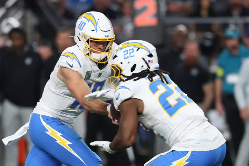 Ein Footballspieler im weißen Chargers-Trikot mit der Nummer 10 übergibt den Ball während eines Spiels an seinen Teamkollegen Najee Harris. Beide tragen Helme und sind fokussiert, mit unscharfen Zuschauern und einer digitalen Nummernanzeige im Hintergrund. Diese Beschreibung wurde mit der FootballR KI automatisch generiert.