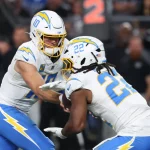 Ein Footballspieler im weißen Chargers-Trikot mit der Nummer 10 übergibt den Ball während eines Spiels an seinen Teamkollegen Najee Harris. Beide tragen Helme und sind fokussiert, mit unscharfen Zuschauern und einer digitalen Nummernanzeige im Hintergrund. Diese Beschreibung wurde mit der FootballR KI automatisch generiert.