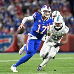 Ein Bills-Footballspieler, Josh Allen, im blauen Trikot mit der Nummer 17, rennt mit dem Ball, während ein Verteidiger der Miami Dolphins, die Nummer 25 in Weiß, ihn auf einem grünen Feld während eines NFL-Spiels mit einer verschwommenen Menschenmenge im Hintergrund verfolgt. Diese Beschreibung wurde mit der FootballR KI automatisch generiert.