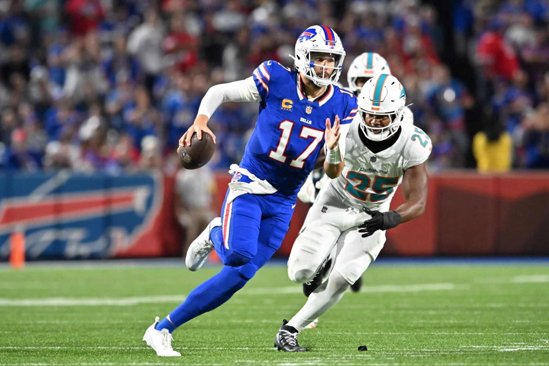 Ein Bills-Footballspieler, Josh Allen, im blauen Trikot mit der Nummer 17, rennt mit dem Ball, während ein Verteidiger der Miami Dolphins, die Nummer 25 in Weiß, ihn auf einem grünen Feld während eines NFL-Spiels mit einer verschwommenen Menschenmenge im Hintergrund verfolgt. Diese Beschreibung wurde mit der FootballR KI automatisch generiert.