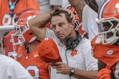 Ein College-Football-Trainer in einem weißen Clemson-Hemd schaut frustriert, hält eine rote Mütze in der Hand und berührt seinen Kopf. Er steht inmitten von Spielern in orangefarbenen Trikots und Helmen, mit einem Headset um den Hals, an der Seitenlinie während der Woche 4. Diese Beschreibung wurde mit der FootballR KI automatisch generiert.