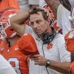 Ein College-Football-Trainer in einem weißen Clemson-Hemd schaut frustriert, hält eine rote Mütze in der Hand und berührt seinen Kopf. Er steht inmitten von Spielern in orangefarbenen Trikots und Helmen, mit einem Headset um den Hals, an der Seitenlinie während der Woche 4. Diese Beschreibung wurde mit der FootballR KI automatisch generiert.