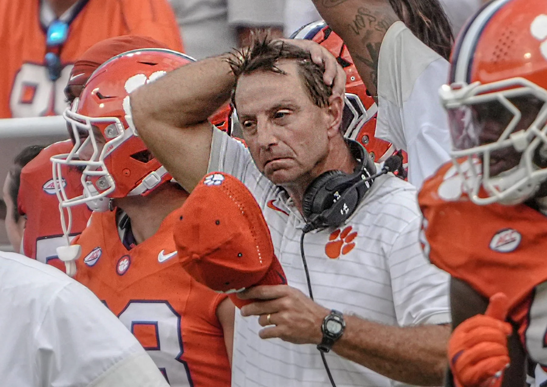 Ein College-Football-Trainer in einem weißen Clemson-Hemd schaut frustriert, hält eine rote Mütze in der Hand und berührt seinen Kopf. Er steht inmitten von Spielern in orangefarbenen Trikots und Helmen, mit einem Headset um den Hals, an der Seitenlinie während der Woche 4. Diese Beschreibung wurde mit der FootballR KI automatisch generiert.