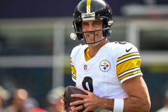 Aaron Rodgers mit schwarzem Helm und weißem Pittsburgh Steelers-Trikot mit der Nummer 8 hält einen Football und lächelt tagsüber auf dem Spielfeld - und erinnert an das Selbstvertrauen von Legenden wie Brett Favre nach denkwürdigen Touchdown-Pässen. Diese Beschreibung wurde mit der FootballR KI automatisch generiert.