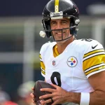 Aaron Rodgers mit schwarzem Helm und weißem Pittsburgh Steelers-Trikot mit der Nummer 8 hält einen Football und lächelt tagsüber auf dem Spielfeld - und erinnert an das Selbstvertrauen von Legenden wie Brett Favre nach denkwürdigen Touchdown-Pässen. Diese Beschreibung wurde mit der FootballR KI automatisch generiert.