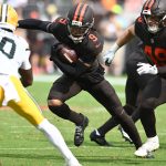 Ein Footballspieler der Browns in einem dunkelbraunen Trikot mit orangefarbenen Streifen läuft mit dem Ball und weicht einem weiß-gelb gekleideten Verteidiger der Packers aus. In der Nähe blockt sein Teamkollege Szmyt auf dem sonnigen, grünen Footballfeld. Diese Beschreibung wurde mit der FootballR KI automatisch generiert.