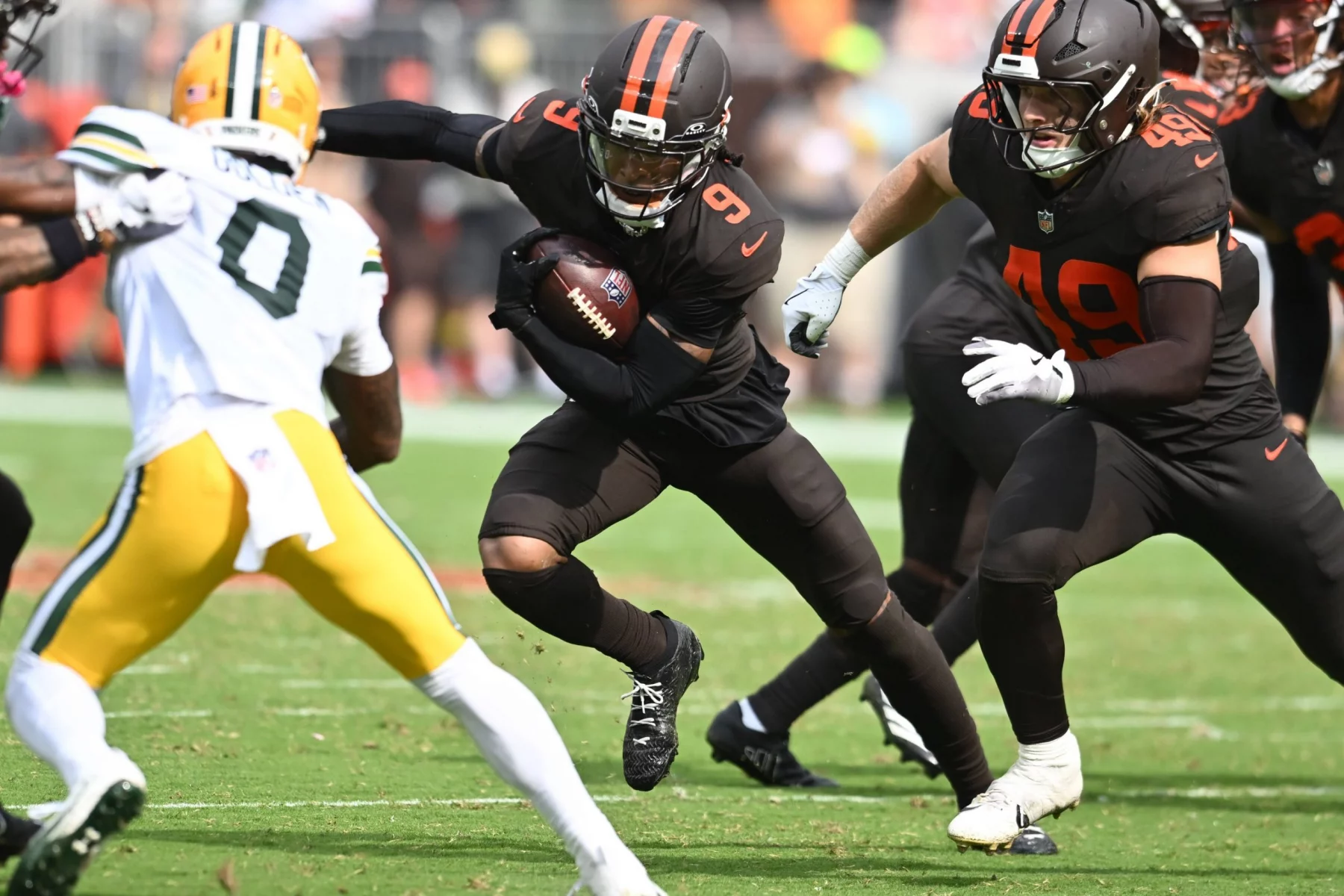 Ein Footballspieler der Browns in einem dunkelbraunen Trikot mit orangefarbenen Streifen läuft mit dem Ball und weicht einem weiß-gelb gekleideten Verteidiger der Packers aus. In der Nähe blockt sein Teamkollege Szmyt auf dem sonnigen, grünen Footballfeld. Diese Beschreibung wurde mit der FootballR KI automatisch generiert.
