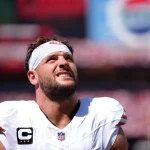 Nick Bosa in einem weißen Trikot mit roten Akzenten, mit Kapitänsabzeichen und weißem Stirnband blinzelt in die Sonne. Die verschwommene Stadionmenge und die Beschilderung deuten auf die Intensität hin - Nick Bosa schaut nach seiner von Kreuzbandriss beendeten Saison zu. Diese Beschreibung wurde mit der FootballR KI automatisch generiert.