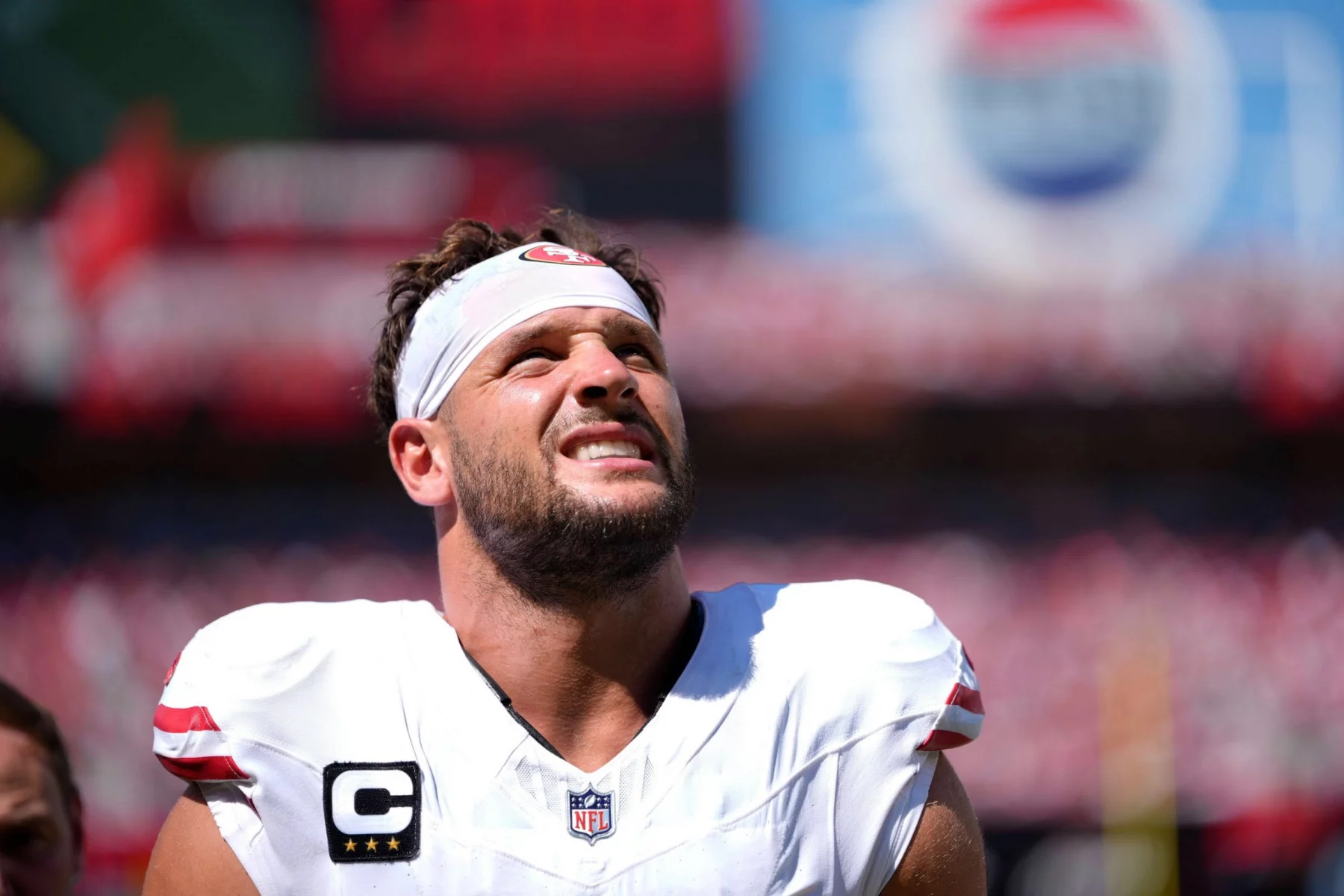 Nick Bosa in einem weißen Trikot mit roten Akzenten, mit Kapitänsabzeichen und weißem Stirnband blinzelt in die Sonne. Die verschwommene Stadionmenge und die Beschilderung deuten auf die Intensität hin - Nick Bosa schaut nach seiner von Kreuzbandriss beendeten Saison zu. Diese Beschreibung wurde mit der FootballR KI automatisch generiert.