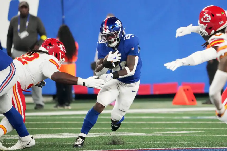Ein Footballspieler in einem blauen Trikot, das an die Giants erinnert, läuft mit dem Ball und weicht zwei gegnerischen Spielern in Rot und Weiß auf einem grünen Feld aus. Die Aktion ist intensiv, da er die Konzentration von Tyrone Tracy Jr. übernimmt, während er ihren Tackles ausweicht. Diese Beschreibung wurde mit der FootballR KI automatisch generiert.