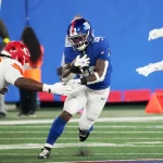 Ein Footballspieler in einem blauen Trikot, das an die Giants erinnert, läuft mit dem Ball und weicht zwei gegnerischen Spielern in Rot und Weiß auf einem grünen Feld aus. Die Aktion ist intensiv, da er die Konzentration von Tyrone Tracy Jr. übernimmt, während er ihren Tackles ausweicht. Diese Beschreibung wurde mit der FootballR KI automatisch generiert.