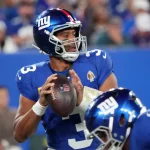 Ein Footballspieler in einer blauen New York Giants-Uniform mit Helm hält einen Football und bereitet sich darauf vor, wie Russell Wilson zu werfen. Er trägt die Nummer 3 und ein Kapitänsabzeichen. Andere Spieler sind zu sehen, und im Hintergrund ist eine unscharfe Stadionmenge zu sehen. Diese Beschreibung wurde mit der FootballR KI automatisch generiert.