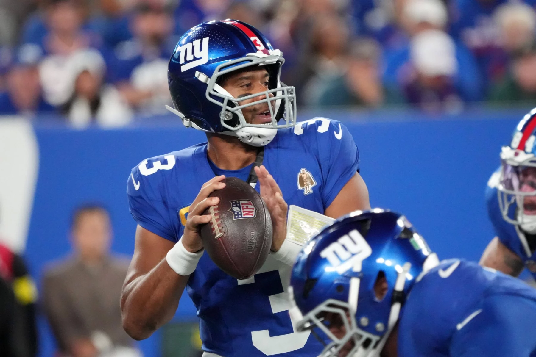 Ein Footballspieler in einer blauen New York Giants-Uniform mit Helm hält einen Football und bereitet sich darauf vor, wie Russell Wilson zu werfen. Er trägt die Nummer 3 und ein Kapitänsabzeichen. Andere Spieler sind zu sehen, und im Hintergrund ist eine unscharfe Stadionmenge zu sehen. Diese Beschreibung wurde mit der FootballR KI automatisch generiert.