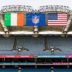 In einem Stadion mit überwiegend leeren blauen Sitzen sind drei große Banner zu sehen: die irische Flagge, das NFL-Logo und die US-Flagge. Digitale Schilder werben für das "GAA Museum & Stadium Tour", was auf die Spannung eines NFL-Spiels in Dublin oder sogar des ersten NFL-Spiels dort hindeutet. Diese Beschreibung wurde mit der FootballR KI automatisch generiert.