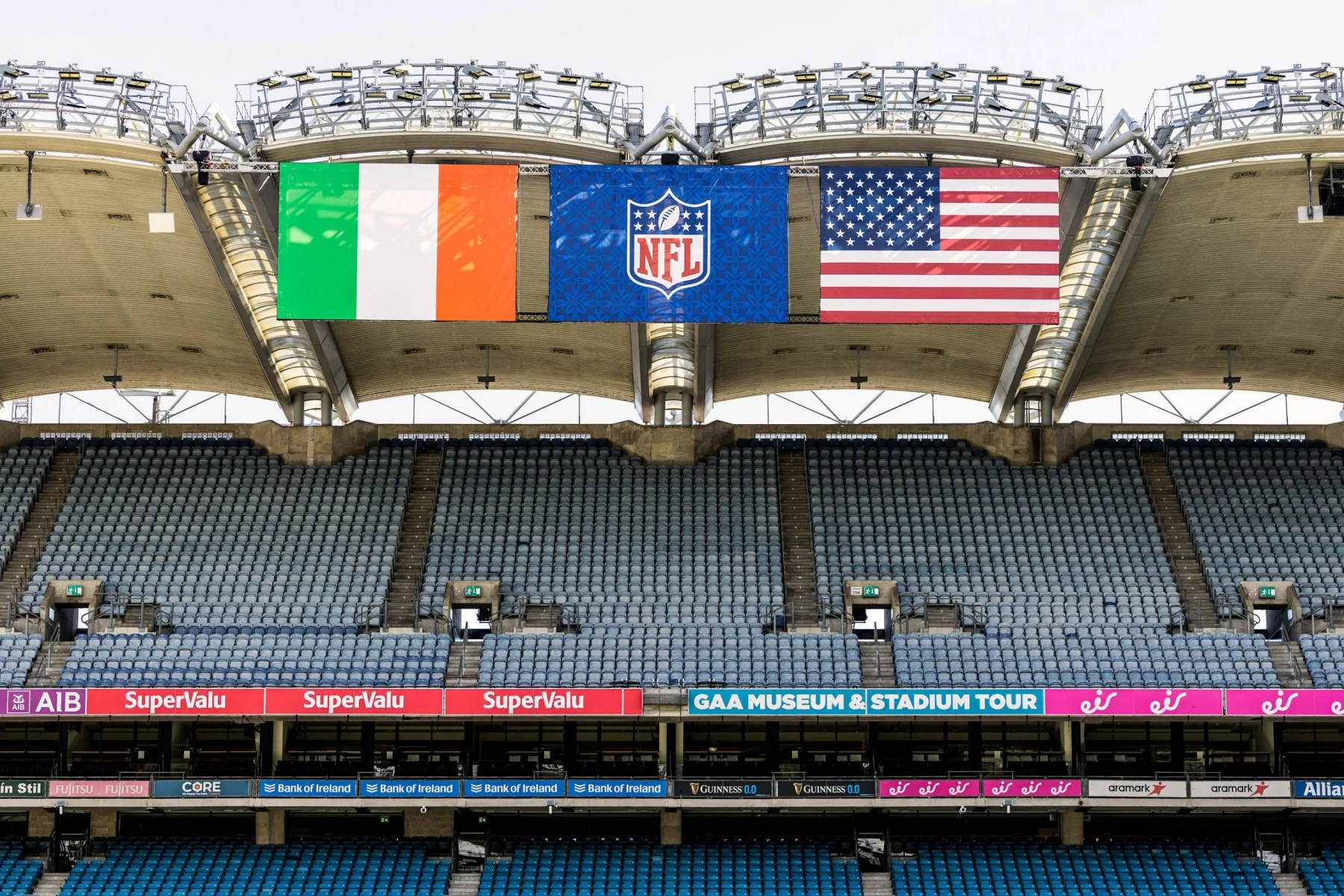 In einem Stadion mit überwiegend leeren blauen Sitzen sind drei große Banner zu sehen: die irische Flagge, das NFL-Logo und die US-Flagge. Digitale Schilder werben für das "GAA Museum & Stadium Tour", was auf die Spannung eines NFL-Spiels in Dublin oder sogar des ersten NFL-Spiels dort hindeutet. Diese Beschreibung wurde mit der FootballR KI automatisch generiert.