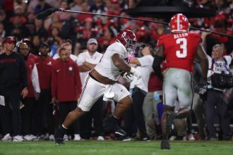 Ein Football-Spieler in einem weißen Alabama-Trikot läuft mit dem Ball, während sich ein Verteidiger von Georgia, Nummer 3, nähert. Trainer und Betreuer schauen bei diesem Woche 5 College Football Rückblick im hellen Stadionlicht von der Seitenlinie aus zu. Diese Beschreibung wurde mit der FootballR KI automatisch generiert.