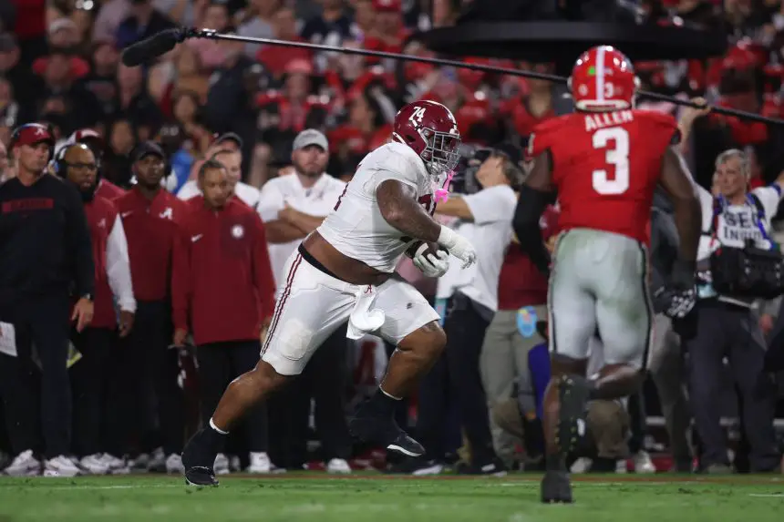 Ein Football-Spieler in einem weißen Alabama-Trikot läuft mit dem Ball, während sich ein Verteidiger von Georgia, Nummer 3, nähert. Trainer und Betreuer schauen bei diesem Woche 5 College Football Rückblick im hellen Stadionlicht von der Seitenlinie aus zu. Diese Beschreibung wurde mit der FootballR KI automatisch generiert.