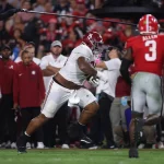 Ein Football-Spieler in einem weißen Alabama-Trikot läuft mit dem Ball, während sich ein Verteidiger von Georgia, Nummer 3, nähert. Trainer und Betreuer schauen bei diesem Woche 5 College Football Rückblick im hellen Stadionlicht von der Seitenlinie aus zu. Diese Beschreibung wurde mit der FootballR KI automatisch generiert.