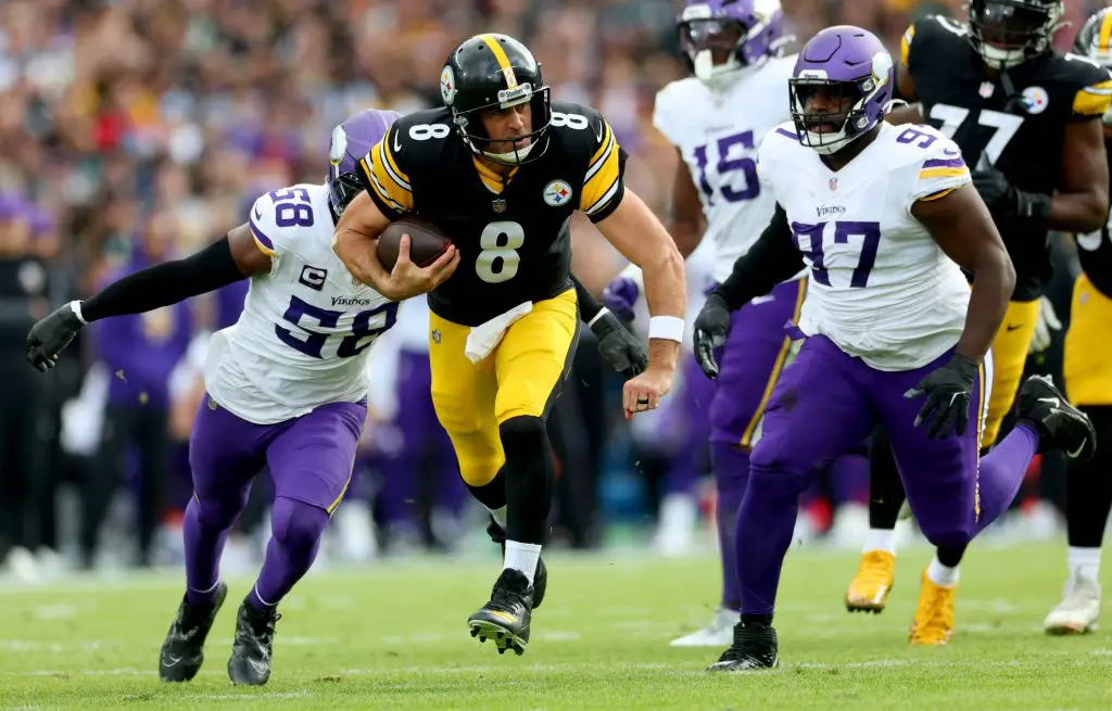 Ein Spieler der Steelers (Nr. 8) läuft mit dem Football, als zwei Verteidiger der Minnesota Vikings in weiß-lila Uniformen versuchen, ihn auf einem Rasenplatz während eines internationalen Spiels zu stoppen. Die Zuschauer sind im Hintergrund unscharf zu erkennen. Diese Beschreibung wurde mit der FootballR KI automatisch generiert.