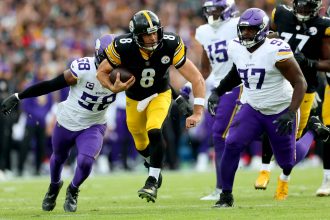 Ein Spieler der Steelers (Nr. 8) läuft mit dem Football, als zwei Verteidiger der Minnesota Vikings in weiß-lila Uniformen versuchen, ihn auf einem Rasenplatz während eines internationalen Spiels zu stoppen. Die Zuschauer sind im Hintergrund unscharf zu erkennen. Diese Beschreibung wurde mit der FootballR KI automatisch generiert.