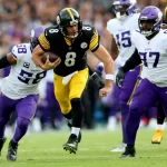 Ein Spieler der Steelers (Nr. 8) läuft mit dem Football, als zwei Verteidiger der Minnesota Vikings in weiß-lila Uniformen versuchen, ihn auf einem Rasenplatz während eines internationalen Spiels zu stoppen. Die Zuschauer sind im Hintergrund unscharf zu erkennen. Diese Beschreibung wurde mit der FootballR KI automatisch generiert.