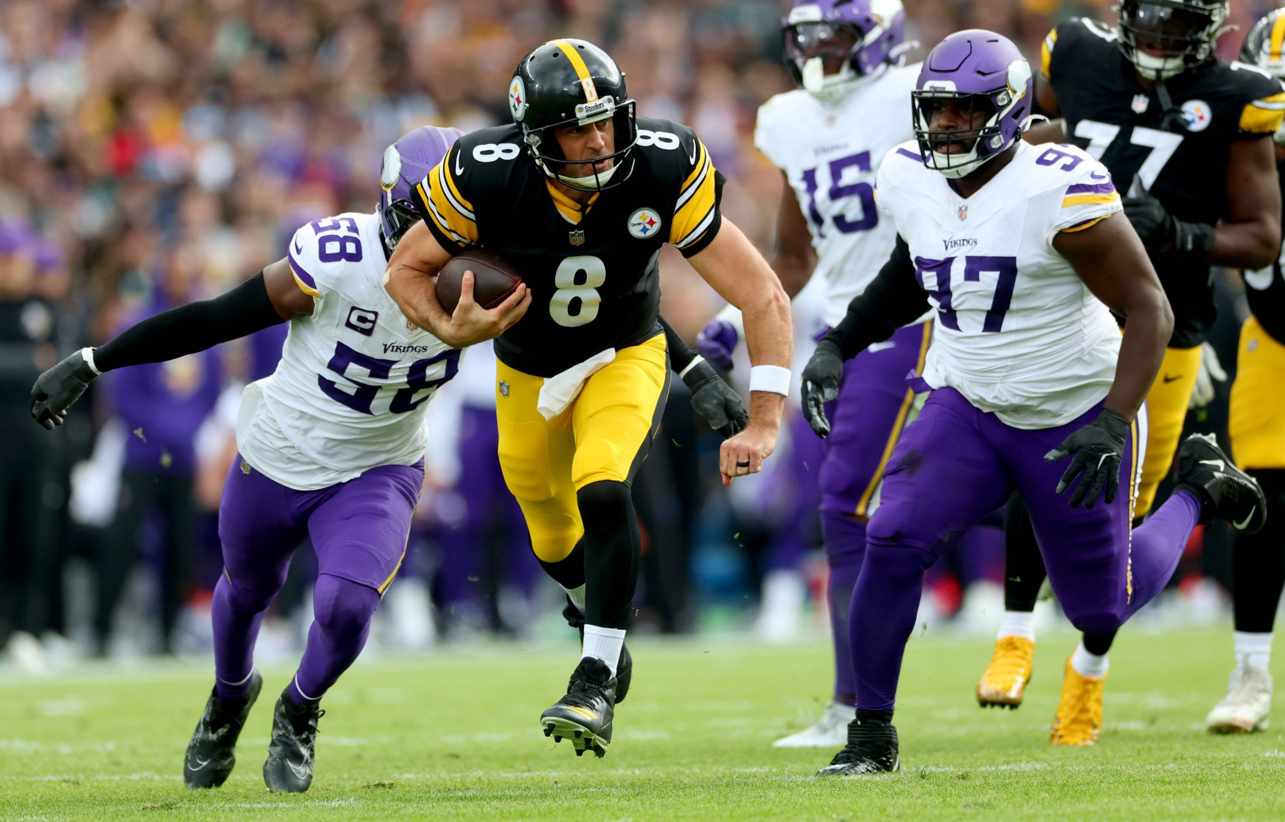 Ein Spieler der Steelers (Nr. 8) läuft mit dem Football, als zwei Verteidiger der Minnesota Vikings in weiß-lila Uniformen versuchen, ihn auf einem Rasenplatz während eines internationalen Spiels zu stoppen. Die Zuschauer sind im Hintergrund unscharf zu erkennen. Diese Beschreibung wurde mit der FootballR KI automatisch generiert.