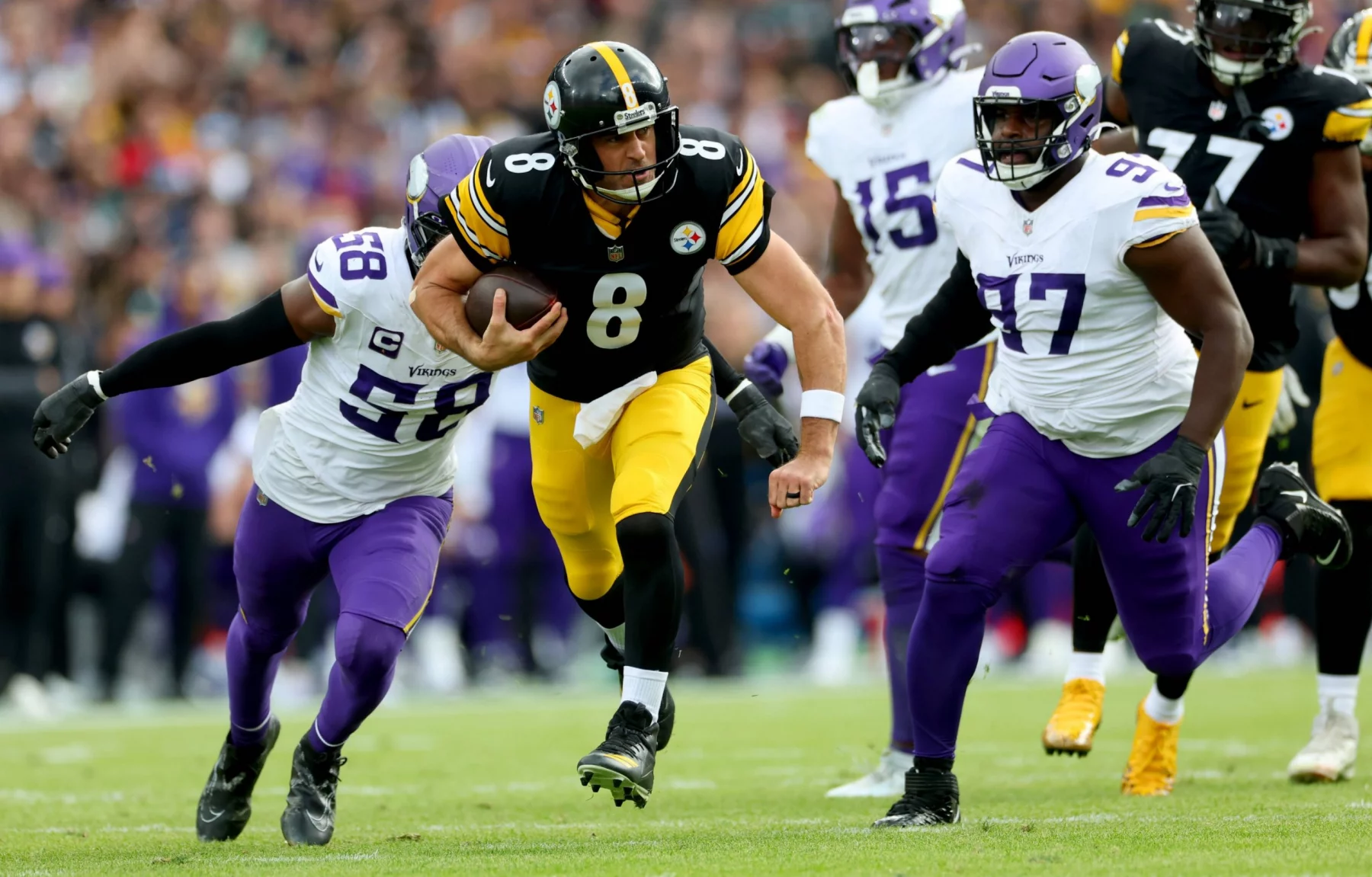 Ein Spieler der Steelers (Nr. 8) läuft mit dem Football, als zwei Verteidiger der Minnesota Vikings in weiß-lila Uniformen versuchen, ihn auf einem Rasenplatz während eines internationalen Spiels zu stoppen. Die Zuschauer sind im Hintergrund unscharf zu erkennen. Diese Beschreibung wurde mit der FootballR KI automatisch generiert.