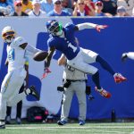 Der blau gekleidete Giants-Receiver Malik Nabers springt mit ausgestrecktem Arm zur Seite, um einen Pass zu fangen, während ein Spieler der Los Angeles Chargers auf dem sonnenbeschienenen, mit Fans gefüllten Spielfeld in der Luft abwehrt. Diese Beschreibung wurde mit der FootballR KI automatisch generiert.