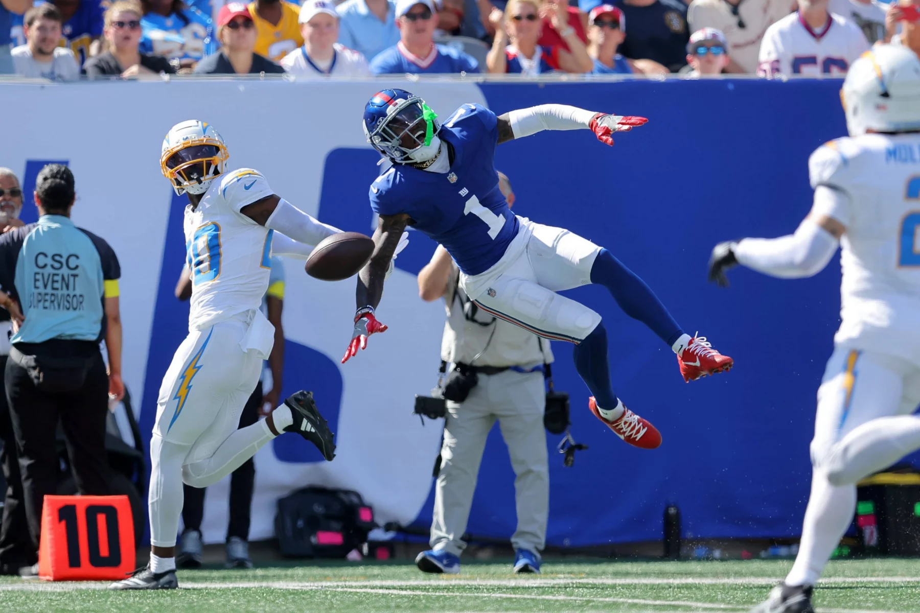 Der blau gekleidete Giants-Receiver Malik Nabers springt mit ausgestrecktem Arm zur Seite, um einen Pass zu fangen, während ein Spieler der Los Angeles Chargers auf dem sonnenbeschienenen, mit Fans gefüllten Spielfeld in der Luft abwehrt. Diese Beschreibung wurde mit der FootballR KI automatisch generiert.